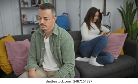 A contemplative man and a distracted woman sit separately on a modern sofa in a cozy, well-decorated living room, suggesting tension. - Powered by Shutterstock