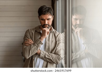 A contemplative man leaning on a large window pane, with his reflection visible. He appears deep in thought, resting his chin on his hand. His expression is one of serious contemplation or concern - Powered by Shutterstock