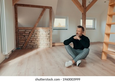 Contemplative man sitting in an empty attic on the wooden floor staring out of the window with a farway expression and chin on hands
