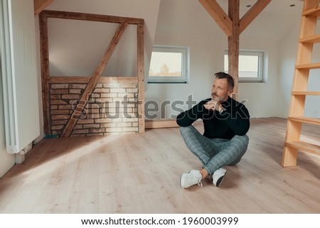 Contemplative man sitting in an empty attic on the wooden floor staring out of the window with a farway expression and chin on hands