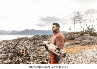 A contemplative man stands by the sea, holding driftwood, lost in the coastal ambience and thoughts. - Powered by Shutterstock