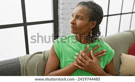 A contemplative mature african american woman in a green shirt relaxes in a bright living room.