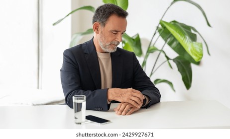 Contemplative mature businessman sitting at a white table with his smartphone, looking away thoughtfully with a glass of water beside him, in a serene environment with greenery - Powered by Shutterstock