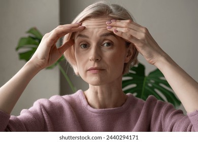 A contemplative mature woman is captured indoors while gently touching her forehead with both hands, appearing introspective or concerned. She wears a soft pink sweater, and her expression conveys - Powered by Shutterstock