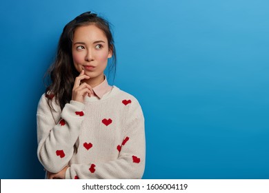 Contemplative Pretty Millennial Young Asian Woman Looks Away, Builds Plans In Mind, Has Thoughtful Face Expression, Long Dark Pony Tail, Wears Sweater With Hearts, Isolated Over Blue Background