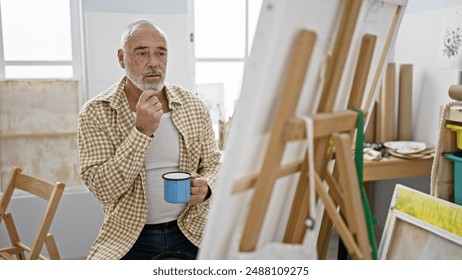 A contemplative senior man with a beard pauses in an art studio, coffee cup in hand, by an easel. - Powered by Shutterstock