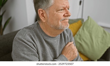 A contemplative senior man sits indoors on a couch, with a neutral expression and casual attire against a cozy home background. - Powered by Shutterstock