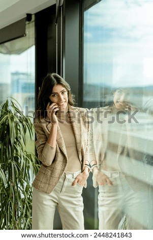 Contemplative woman using mobile phone and gazes out of a window, with a thoughtful expression and city views behind her