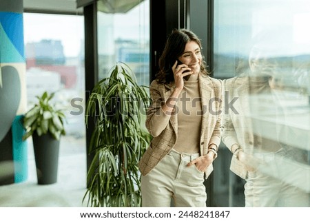 Contemplative woman using mobile phone and gazes out of a window, with a thoughtful expression and city views behind her