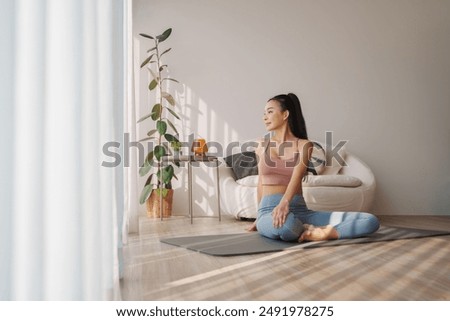 Contemplative young Asian woman in yoga attire seated on a mat in her living room, engaging in a morning stretch routine with soft shadows dancing on the wall