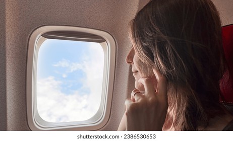 A contemplative young beautiful woman gazes out of an airplane window, lost in thought as she takes in the serene sky and fluffy clouds from above. - Powered by Shutterstock