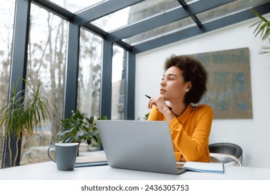 Contemplative young black woman with stylish afro hairstyle gazes out window while working at her laptop in bright, naturally lit modern office space - Powered by Shutterstock