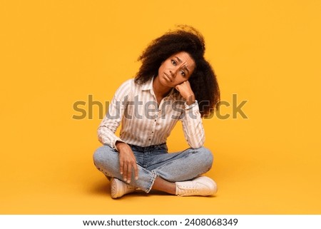 Contemplative young black woman with voluminous curly hair sits cross-legged on floor, hand resting on her cheek, deep in thought against vivid yellow background