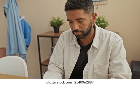 A contemplative young man with a beard sits indoors in a casual room, appearing thoughtful or fatigued. - Powered by Shutterstock