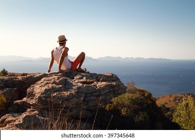 Contemplative young man sitting on the rocks on the top of the mountain at sunset. Looking at distance with the blue sea in front. Wearing a straw at in summer. - Powered by Shutterstock