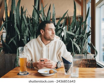 A contemplative young man with a smartphone, enjoying a glass of beer in a cozy cafe setting, surrounded by indoor plants - Powered by Shutterstock