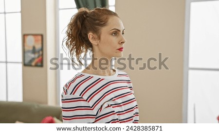 A contemplative young woman indoors wearing a striped shirt with a modern living room backdrop.