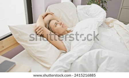 A contemplative young woman lies in bed, wrapped in white sheets in a softly lit bedroom, embodying relaxation and comfort.