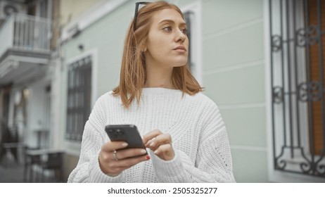 A contemplative young woman in a white sweater using a smartphone on a city street. - Powered by Shutterstock