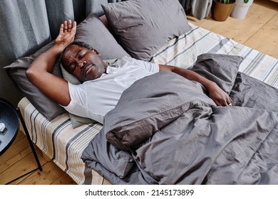 Contemporary African American senior man napping under grey blanket in bed in the morning while keeping ihs head on pillow