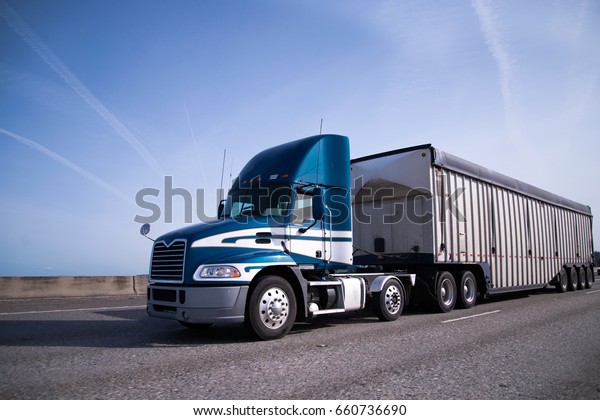 Contemporary big rig blue\
semi truck with high day cab and covered semi trailer for delivery\
bulk cargo moving on the wide highway with blue sky and little\
clouds on\
background