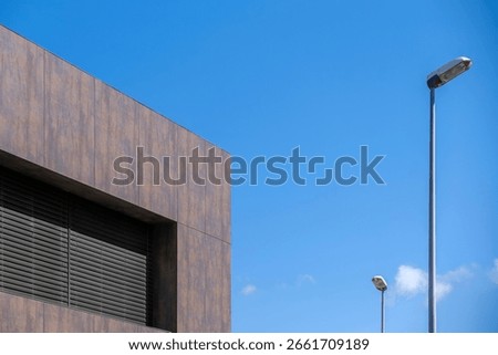 Contemporary Building Facade With Dark Cladding And Streetlights Against Clear Blue Sky. Modern architecture, urban design, geometric shapes, commercial property, exterior view, bright sunny day