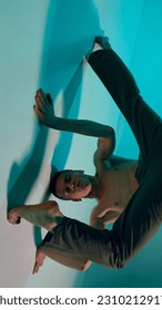 Contemporary dance style. Upside down. Young shirtless man dancing contemp, experimental dance over blue, cyan studio background. Concept of art, body aesthetics, motion, action, inspiration.