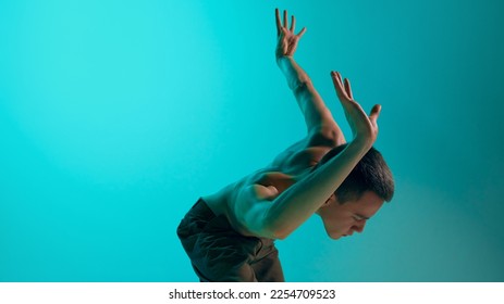 Contemporary dance style. Young shirtless man dancing over blue cyan color studio background. Flexibility of movements. Concept of art, body aesthetics, motion, action, inspiration. - Shutterstock ID 2254709523