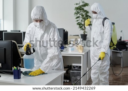Contemporary disinfection service company staff in protective workwear carrying out their work in spacious openspace office