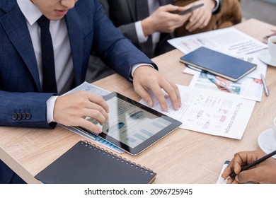Contemporary economist in suit looking through charts and diagrams in tablet while sitting by table among coworkers