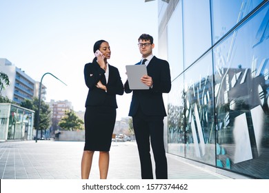 Contemporary Elegant Man Watching Tablet While Standing With Young Female Black Coworker On Street Speaking On Mobile Phone In Back Lit