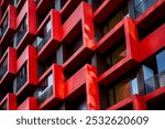 Contemporary facade showcasing a pattern of square-shaped protruding balconies in red and dark gray tones, complemented by glass windows and railings, emphasizing modern urban design and geometry.