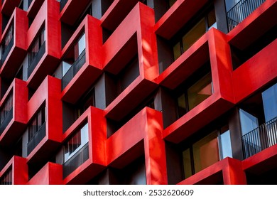 Contemporary facade showcasing a pattern of square-shaped protruding balconies in red and dark gray tones, complemented by glass windows and railings, emphasizing modern urban design and geometry.