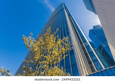 A contemporary glass-clad skyscraper reaches into the clear blue sky, with budding spring trees in the foreground. The perspective captures the contrast between nature and urban architecture - Powered by Shutterstock
