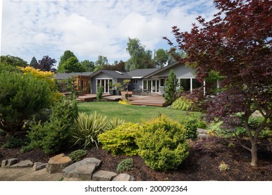 A Contemporary Home Can Be Seen Beyond The Backyard Lawn With Rock And Evergreen Landscaping In The Foreground On A Mild Summer Day In The Pacific Northwest.