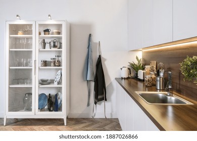 Contemporary home with design interior, white kitchen cupboards, kitchenware in cabinet, empty and clean sink at wooden countertop, apron on wall hook - Powered by Shutterstock