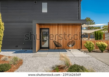contemporary home exterior with black metal siding cedar trim shingles showing front door patio back yard and two black bucket chairs with sunny blue skies