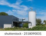 A contemporary industrial building sits beside a tall silo, surrounded by greenery. The structure features a sleek design and is illuminated by bright sunlight, showcasing its architectural details.