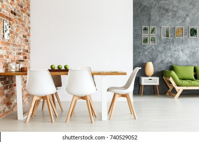 Contemporary Interior Design Of Dining Space With Communal Table Against Brick Wall And Open Living Room With Simple Sofa And Plant Decorations On Textured Gray Wall