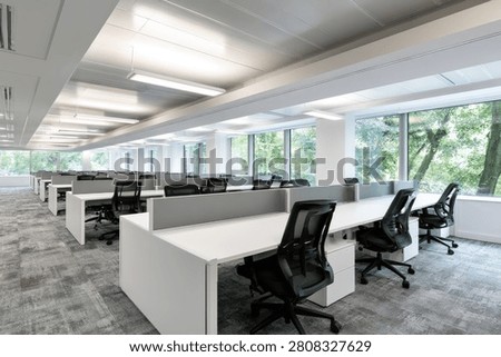 A contemporary office interior featuring rows of workstations, ergonomic chairs, and large windows offering a view of trees.