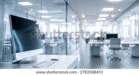 A contemporary office space featuring a computer on a desk in the foreground, with blurred workstations and glass partitions in the background.