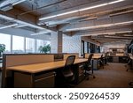 A contemporary office space featuring an open-plan layout with rows of wooden desks and ergonomic chairs. The workspace is well-lit with ceiling lights and natural light from large windows. 