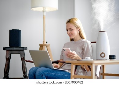 Contemporary smart humidifier on table emitting water vapor and moisturizing air in cozy room where adorable caucasian lady is working on laptop, enjoy spare time at home, in smart house