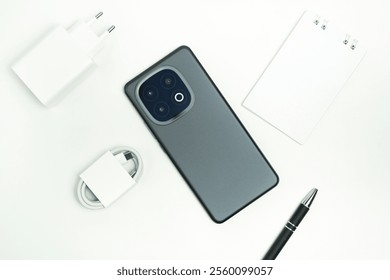 Contemporary smartphone and complementary items, including a charging adapter, USB cable, pen, and notebook, arranged on a minimalistic white surface.