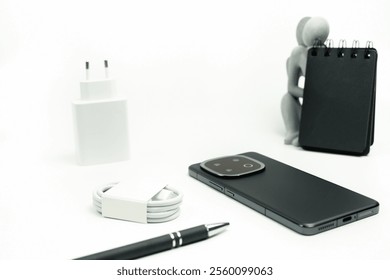 Contemporary smartphone and complementary items, including a charging adapter, USB cable, pen, and notebook, arranged on a minimalistic white surface.