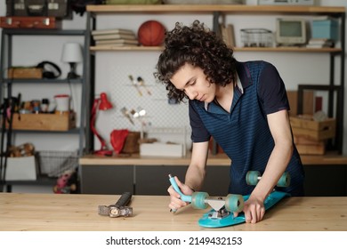 Contemporary teenage guy fitting wheels of skateboard while bending over wooden table or workbench in garage of his house