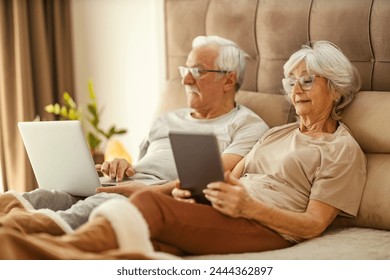 Contemporary trendy senior couple sitting on a bed at cozy bedroom and using a laptop and tablet fro entertainment. - Powered by Shutterstock