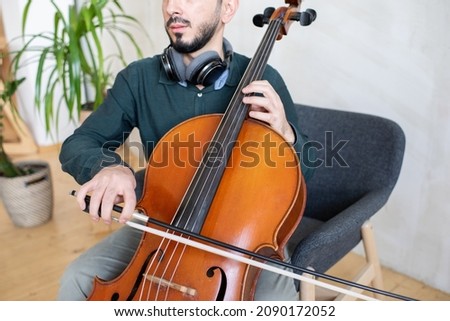 Contemporary young man gliding fiddlestick against strings of cello while sitting in armchair during lesson or repetition