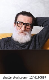 Contemporay senior bearded man in glasses and grey pullover using laptop and browsing internet while sitting on the yellow sofa in the light living room, modern technology, communication concept