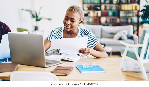 Content African American businesswoman in casual outfit sitting at table with netbook and doing paperwork while working remotely in cozy home office